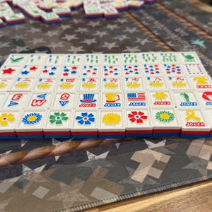 May include: A set of white Mahjong tiles with red, blue, and yellow accents. The tiles feature various symbols, numbers, and characters, including flowers, stars, and the word "Joker." The tiles are arranged on a patterned surface.