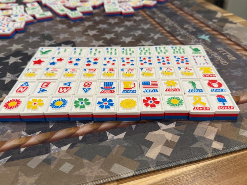 May include: A set of white Mahjong tiles with red, blue, and yellow accents. The tiles feature various symbols, numbers, and characters, including flowers, stars, and the word "Joker." The tiles are arranged on a patterned surface.