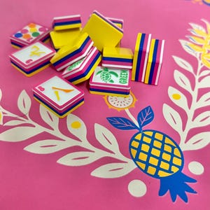 May include: A collection of colorful game tiles with yellow, pink, and blue accents. Some tiles display numbers and a turtle graphic with the word "GREEN". The tiles are arranged on a pink surface with a pineapple and floral design.