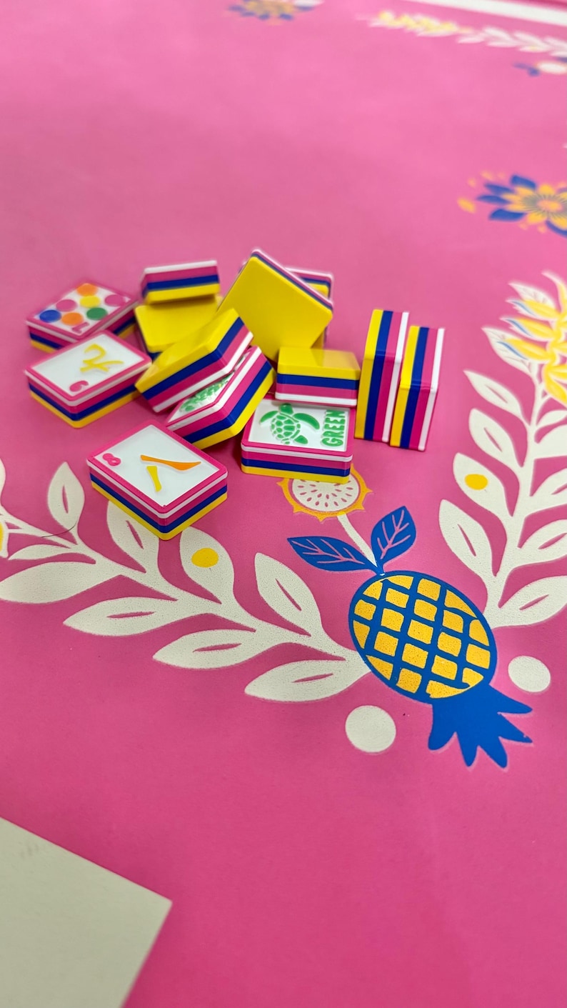 May include: A collection of colorful game tiles with yellow, pink, and blue accents. Some tiles display numbers and a turtle graphic with the word "GREEN". The tiles are arranged on a pink surface with a pineapple and floral design.