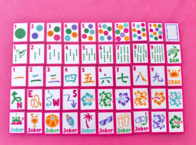 May include: A full Mahjong set with pink edging and white tiles. The tiles display diverse symbols, numbers, and Chinese characters. Some tiles have the word "Joker" at the bottom. The set is laid out on a pink surface.