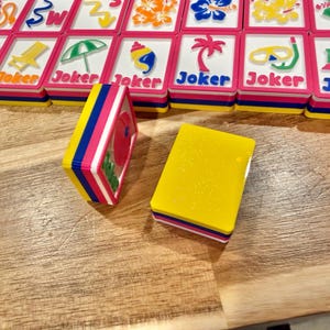 May include: A set of colorful Mahjong tiles with a tropical theme. The tiles feature images of flowers, beach chairs, and palm trees. The tiles are stacked on a wooden surface. The word "Joker" is printed on some tiles.