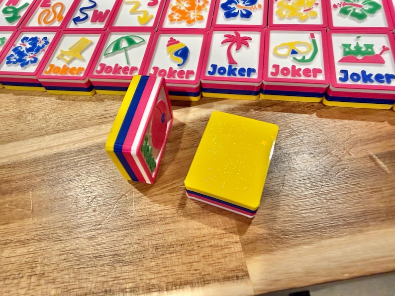 May include: A set of colorful Mahjong tiles with a tropical theme. The tiles feature images of flowers, beach chairs, and palm trees. The tiles are stacked on a wooden surface. The word "Joker" is printed on some tiles.