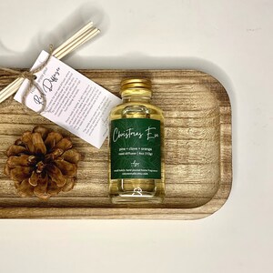 May include: A clear glass bottle with a gold lid filled with a yellow liquid. The bottle is labeled "Christmas Eve" with the scent "pine, clove & orange" and is a 4 oz reed diffuser. The bottle is sitting on a wooden tray with a pine cone.