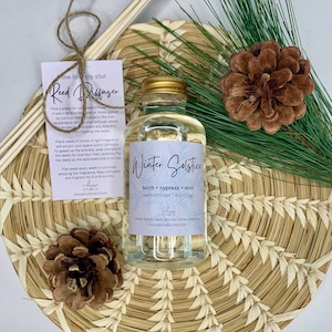 May include: A clear glass bottle with a gold lid filled with a light-colored liquid. The bottle is labeled "Winter Solstice" and lists the scent as "birch + cypress + mint" reed diffuser. The bottle is sitting on a woven straw mat with a pine branch and pine cone.