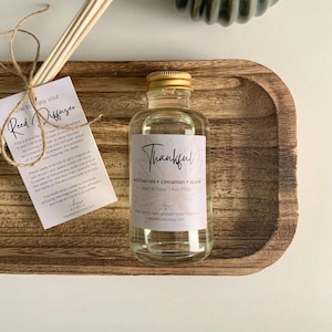May include: A clear glass bottle with a gold lid filled with a reed diffuser. The label is white with black text that reads "Thankful" and "wild berries + cinnamon + apple reed diffuser | 4oz (113g)". The bottle is sitting on a wooden tray with a set of wooden reeds.