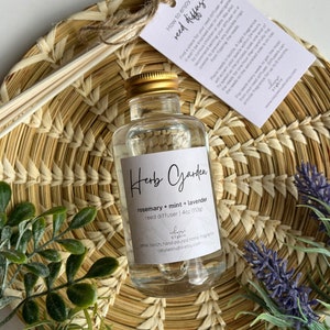 May include: A clear glass bottle with a gold lid filled with a reed diffuser oil. The bottle label reads "Herb Garden rosemary + mint + lavender reed diffuser 4oz (113g)" and "small batch, hand poured home fragrance celysestudio.etsy.com".