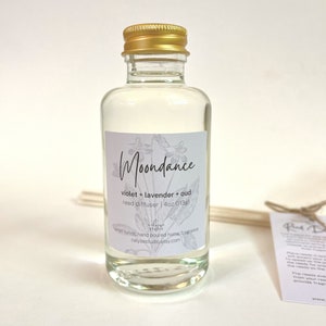 May include: A clear glass bottle with a gold lid filled with a clear liquid. The bottle has a white label with the text "Moondance", "violet + lavender + oud", "reed diffuser | 4oz (113g)", "small batch hand poured home fragrance", and "celysestudio.etsy.com".