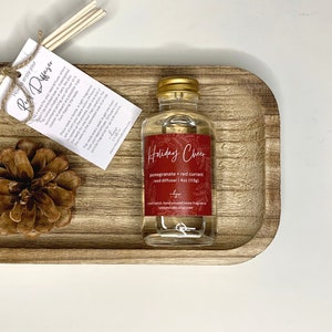 May include: A clear glass bottle with a gold lid filled with a red currant and pomegranate scented reed diffuser. The bottle has a red label with the text "Holiday Cheer" and "pomegranate + red currant reed diffuser | 4oz (113g)". The bottle is sitting on a wooden tray with a pine cone.