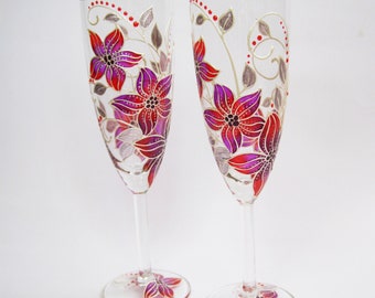 May include: Two clear champagne flutes with hand-painted floral designs. The flowers are red and purple with gold accents and gray leaves. The stems and bases are clear, with a floral design on the base.