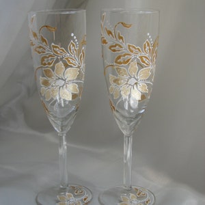 May include: Two champagne flutes with hand-painted gold and white floral designs. The glasses are clear and have a delicate stem.