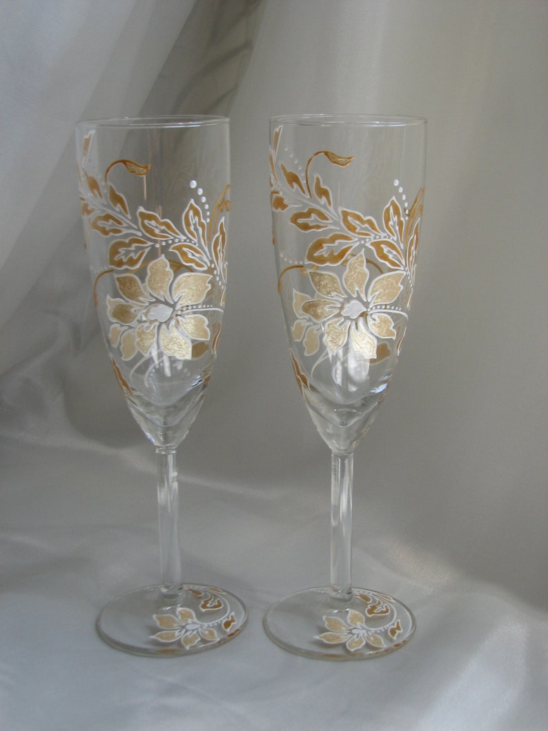 May include: Two champagne flutes with hand-painted gold and white floral designs. The glasses are clear and have a delicate stem.