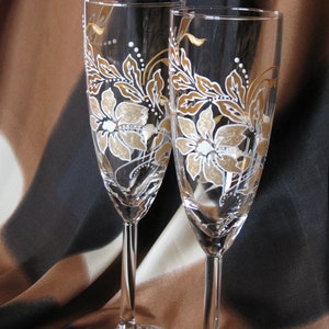 May include: Two champagne flutes with gold and white floral designs painted on the glass. The flutes are on a brown and black patterned fabric background.