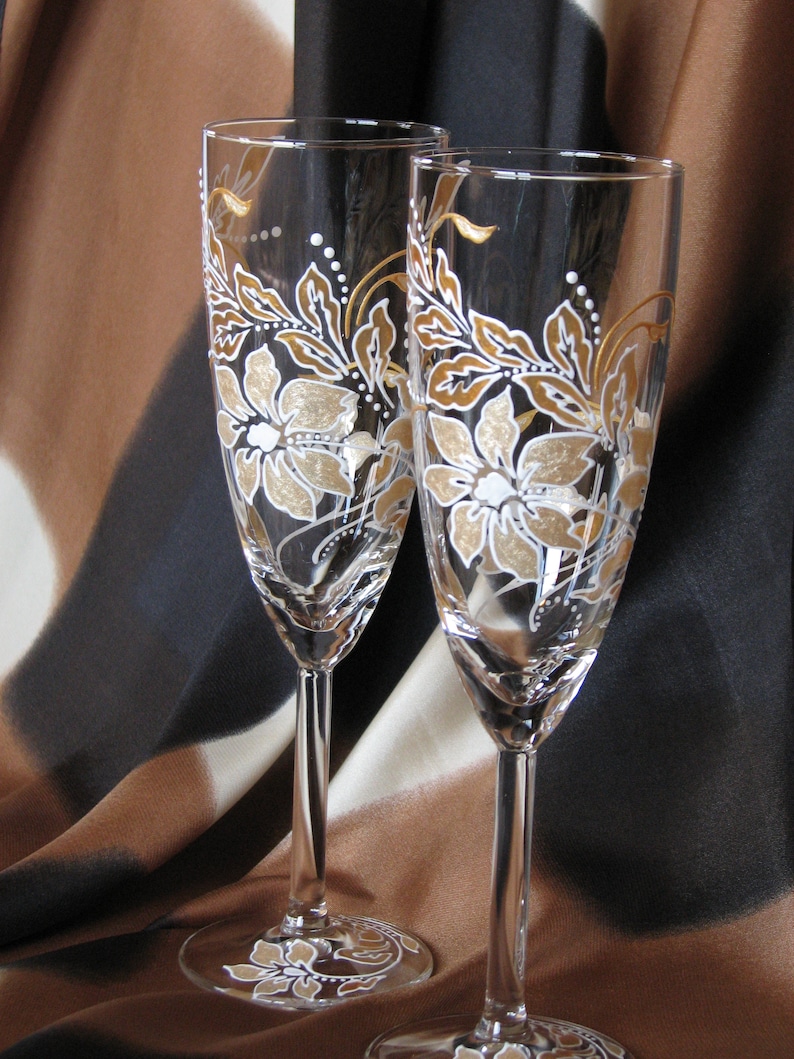 May include: Two champagne flutes with gold and white floral designs painted on the glass. The flutes are on a brown and black patterned fabric background.