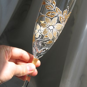 May include: A clear glass champagne flute with a hand-painted design of white and gold flowers and leaves.