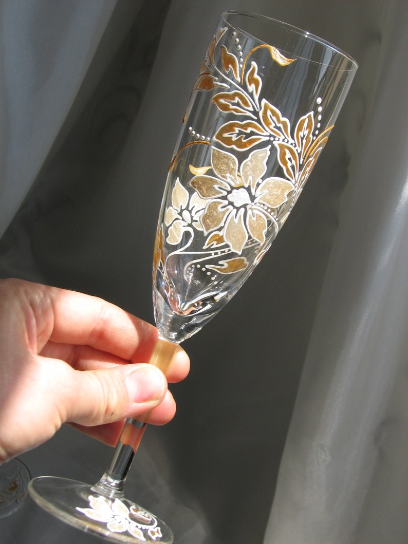 May include: A clear glass champagne flute with a hand-painted design of white and gold flowers and leaves.