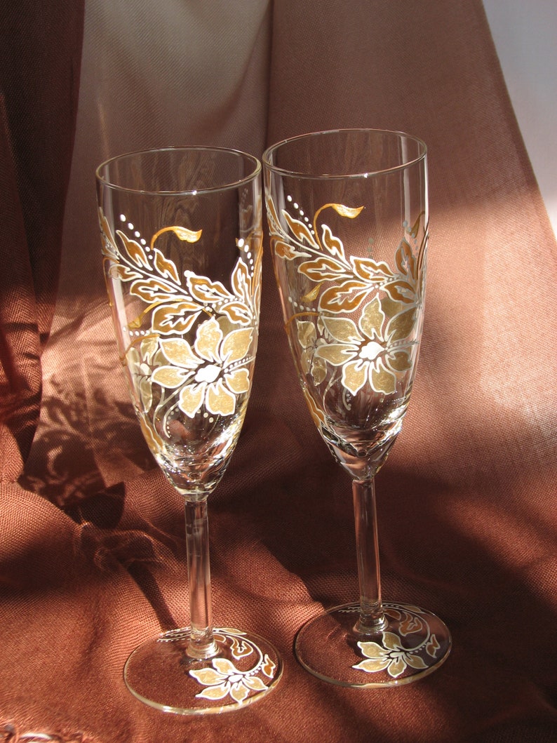 May include: Two champagne flutes with hand-painted floral designs in shades of brown and white. The flowers are stylized and have a vintage feel.
