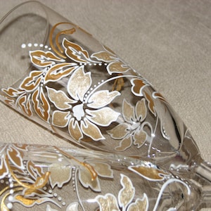 May include: Two champagne flutes with a floral design painted in gold and white. The design features leaves and flowers.