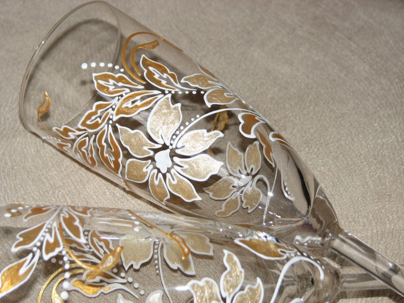 May include: Two champagne flutes with a floral design painted in gold and white. The design features leaves and flowers.