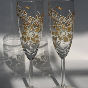May include: Two clear glass champagne flutes with gold and white floral designs. The flutes have a long stem and a wide bowl.