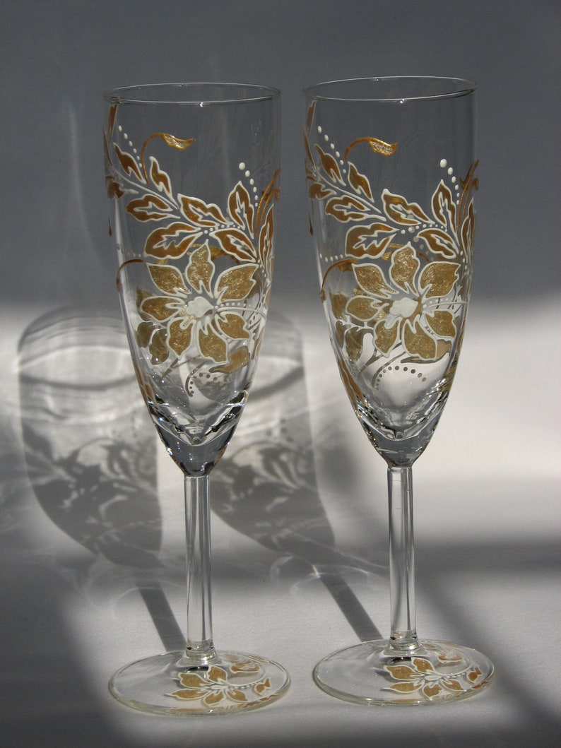 May include: Two clear glass champagne flutes with gold and white floral designs. The flutes have a long stem and a wide bowl.