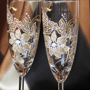 May include: Two clear glass champagne flutes with gold and white floral designs painted on the glass.