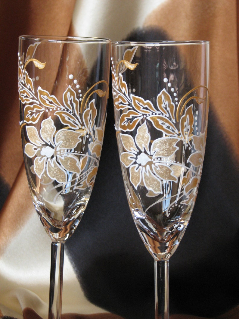 May include: Two clear glass champagne flutes with gold and white floral designs painted on the glass.