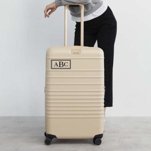 Könnte beinhalten: Beigefarbener Koffer mit einem schwarzen Etikett, auf dem "ABC" steht. Der Koffer hat eine strukturierte Oberfläche und ist auf Rollen.