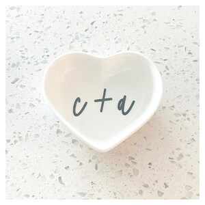 Puede incluir: Plato de cer&aacute;mica blanco en forma de coraz&oacute;n con el texto "c + a" en letras negras.