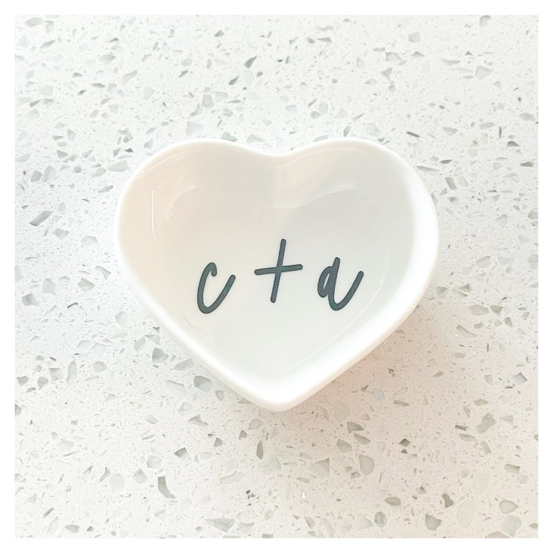 Puede incluir: Plato de cer&aacute;mica blanco en forma de coraz&oacute;n con el texto "c + a" en letras negras.