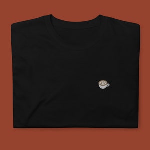 Puede incluir: Camiseta negra con una pequeña imagen bordada de una taza de café blanca con un diseño de arte latte marrón.