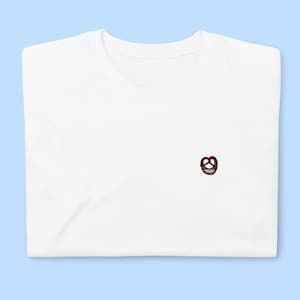 Può includere: Maglietta bianca con un piccolo pretzel marrone ricamato sul davanti.