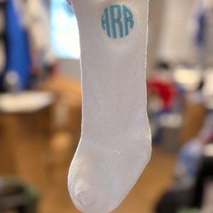 May include: White baby sock with a light blue monogram. The sock is made of a soft material and has a ribbed cuff. The monogram is in a circular design.