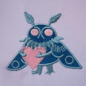 Może przedstawiać: Naszywka haftowana przedstawiająca niebieskiego motyla trzymającego różowe serce. Motyl ma duże, okrągłe oczy i pierzaste czułki.