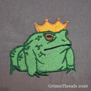 以下が含まれることがあります： 金色の王冠をかぶった緑色のカエルの刺繍パッチ。カエルは詳細なステッチが施されており、灰色の背景に配置されています。「GrimmThreads 2026」の文字が下部にあります。