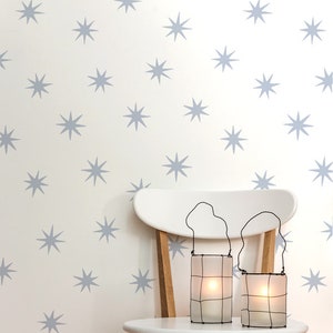 May include: Two white wire-framed candle lanterns with a white candle inside each, sitting on a white wooden chair with a light wood base. The chair is in front of a white wall with a repeating pattern of light blue stars.