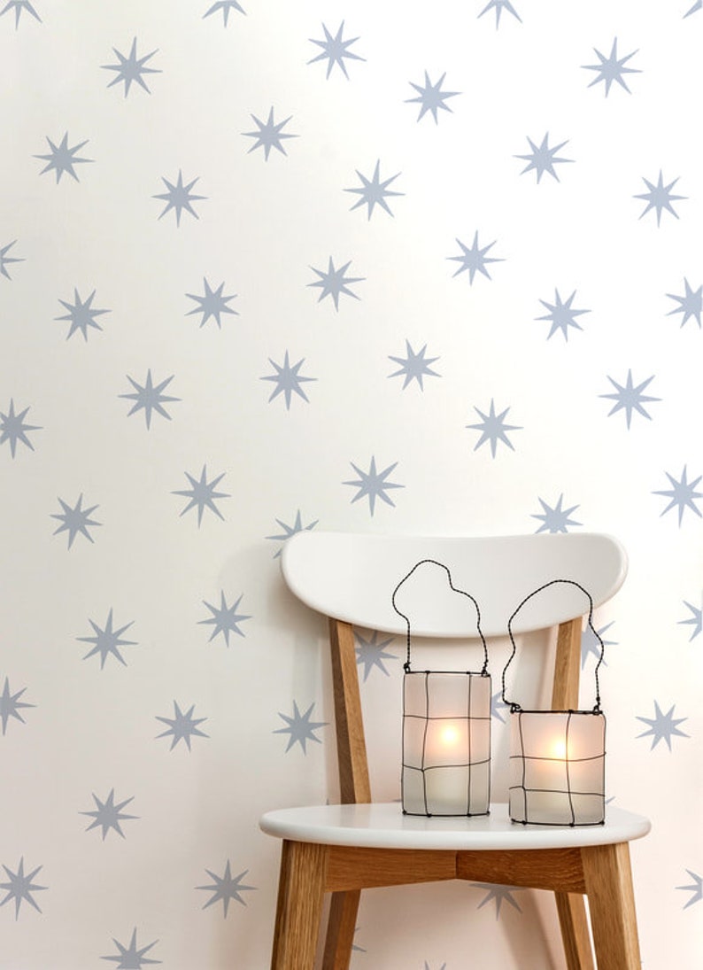 May include: Two white wire-framed candle lanterns with a white candle inside each, sitting on a white wooden chair with a light wood base. The chair is in front of a white wall with a repeating pattern of light blue stars.