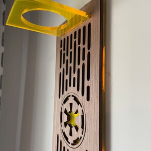 May include: A wooden wall mount with a yellow acrylic shelf and a cutout of the Imperial symbol from Star Wars. The wall mount has a repeating pattern of vertical lines.