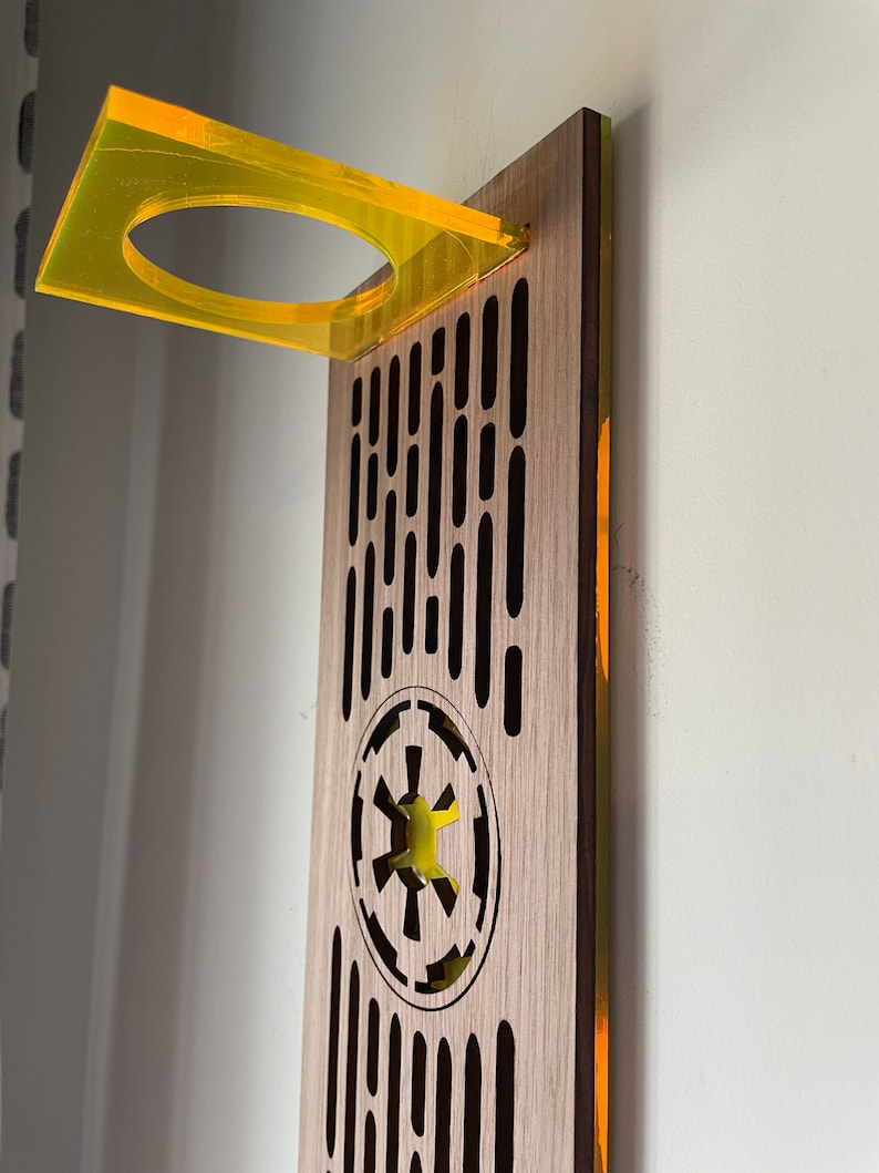 May include: A wooden wall mount with a yellow acrylic shelf and a cutout of the Imperial symbol from Star Wars. The wall mount has a repeating pattern of vertical lines.