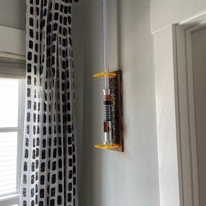 May include: A white lightsaber with a yellow and orange handle is mounted on a wall-mounted display. The display is made of wood and has a clear plastic front. The lightsaber is mounted on a white wall.