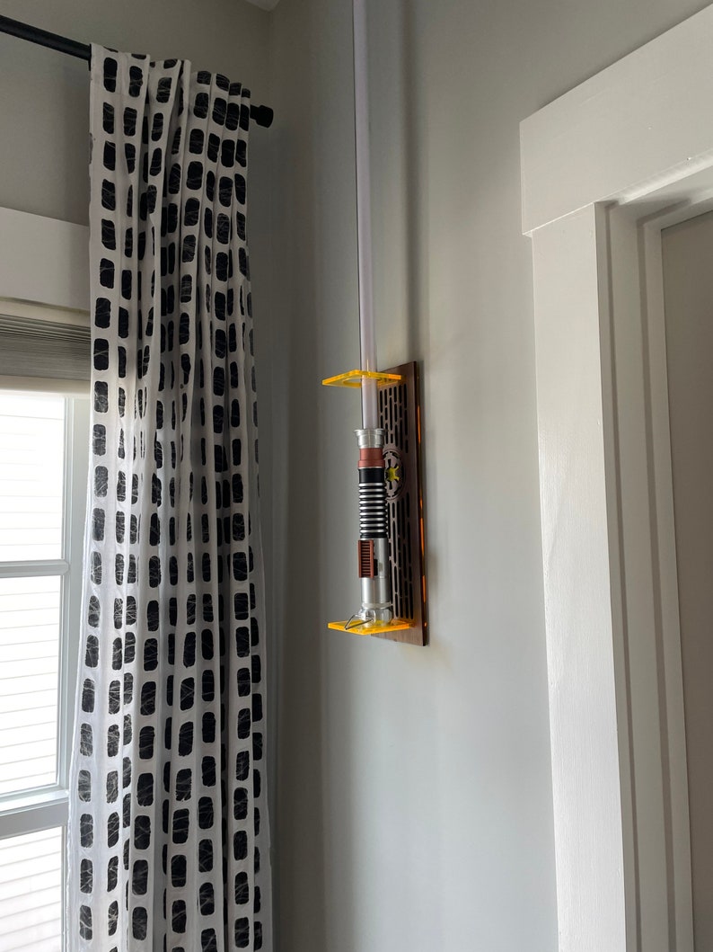 May include: A white lightsaber with a yellow and orange handle is mounted on a wall-mounted display. The display is made of wood and has a clear plastic front. The lightsaber is mounted on a white wall.
