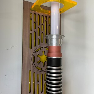 May include: A lightsaber wall mount with a wooden base and a yellow acrylic holder. The lightsaber is silver and black with a copper accent. The base features a yellow Imperial symbol.