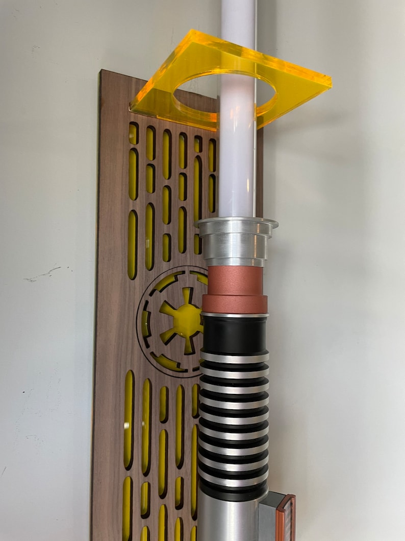 May include: A lightsaber wall mount with a wooden base and a yellow acrylic holder. The lightsaber is silver and black with a copper accent. The base features a yellow Imperial symbol.