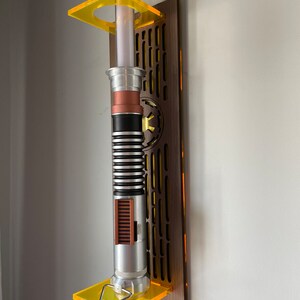 May include: A lightsaber wall mount made of wood with a yellow acrylic stand. The lightsaber is silver with black and brown accents.