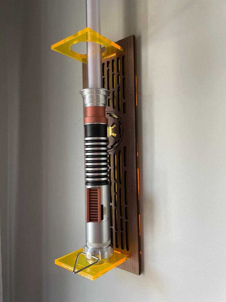May include: A lightsaber wall mount made of wood with a yellow acrylic stand. The lightsaber is silver with black and brown accents.