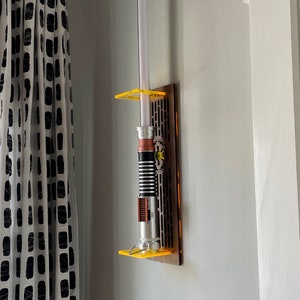 May include: A lightsaber wall mount with a brown wooden base and yellow acrylic accents. The lightsaber is silver and black with a brown handle.