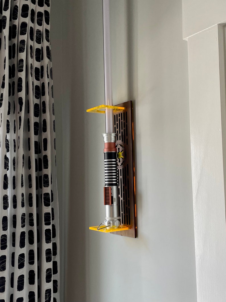 May include: A lightsaber wall mount with a brown wooden base and yellow acrylic accents. The lightsaber is silver and black with a brown handle.