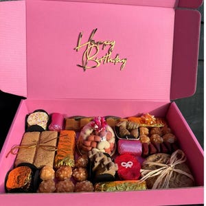 May include: A pink gift box with a gold "Happy Birthday" sign on the lid. The box is filled with a variety of wrapped treats, including chocolate, cookies, and candies.