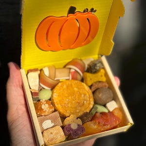 May include: A yellow cardboard box with an orange pumpkin cutout on the lid, filled with a variety of dog treats. The treats include chews, biscuits, and other snacks in various shapes and colors. The box is held open.