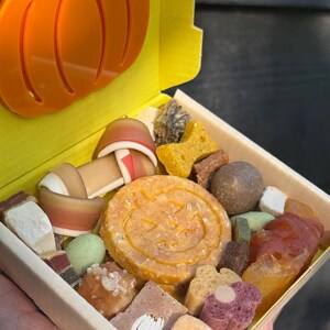 May include: A wooden box filled with assorted dog treats. The treats include a large round biscuit with a jack-o'-lantern face, bone-shaped biscuits, and other colorful, textured snacks. An orange pumpkin charm is attached to the box.